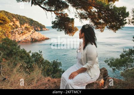 An attractive young lady sat on the edge of a cliff surround by trees looking out at the sea Stock Photo