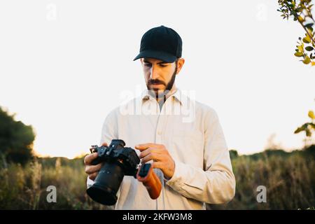 Portrait of a young bearded man, wearing a black cap, with his digital camera at countryside Stock Photo