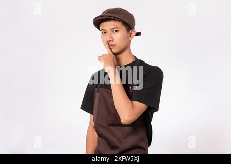 Handsome barista asian man wearing brown apron and black t-shirt isolated over white background asking to be quiet with finger on lips. shut up, secre Stock Photo