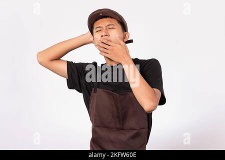 Handsome barista asian man wearing brown apron and black t-shirt isolated over white background yawning covering mouth with hand. sleepy and tired exp Stock Photo