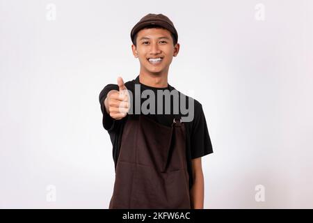Handsome barista asian man wearing brown apron and black t-shirt isolated over white background doing thumbs up gesture with positive expression. Like Stock Photo