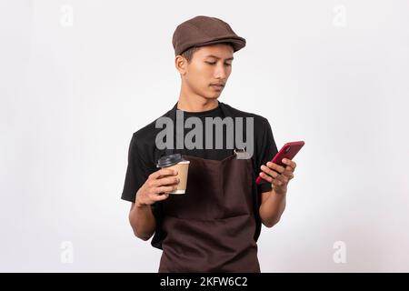 handsome barista asian man wearing brown apron and black t-shirt isolated over white background holding coffee while using mobile phone. concept of ge Stock Photo