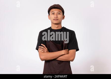 Handsome barista asian man wearing brown apron and black t-shirt isolated over white background standing with crossed arms smiling confident looking a Stock Photo