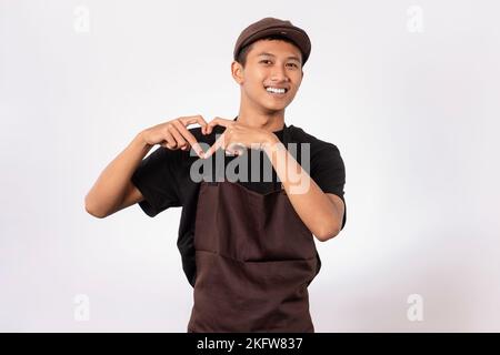 Handsome barista asian man wearing brown apron and black t-shirt isolated over white background smiling doing heart symbol shape with hands. Stock Photo