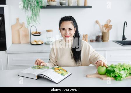 An optimistic young woman looks into the camera and smiles as she sits in her kitchen. Reading a magazine about healthy eating and lifestyle Stock Photo