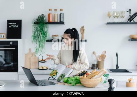 A pretty young woman food blogger is streaming and live-streaming through her laptop and showing in her book how to cook Stock Photo