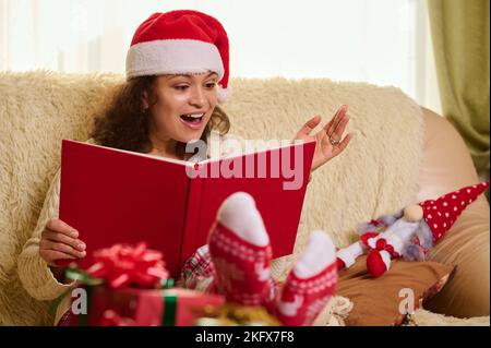 Beautiful Hispanic woman in Santa hat and socks with Christmas pattern, reading fairy tales at winter holidays. New Year Stock Photo