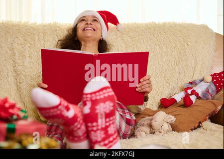 Attractive woman in Santa hat and socks with Christmas pattern, reading fairy tales, resting at home at winter holidays Stock Photo