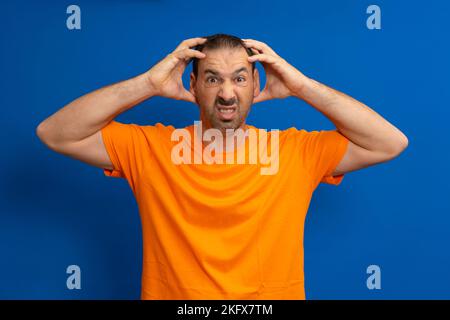 Stressed latin man screaming in panic or anger, shocked, terrified or furious, with hands on head isolated over blue background. Stock Photo