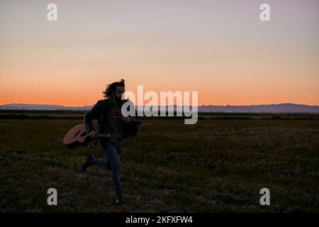 caucasian young man with long hair in fur jacket running through the big lonely field holding a guitar at sunset Stock Photo