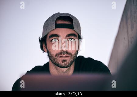face of bearded caucasian guy with nose piercing gray cap black t-shirt sitting illuminated by his laptop screen looking at camera at sunset Stock Photo