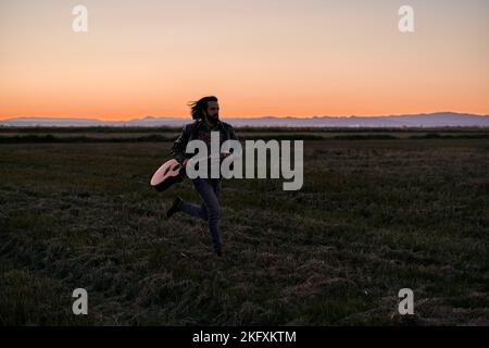 Caucasian young man with long hair in fur jacket running across the plain away from the mountains holding a guitar at sunset Stock Photo