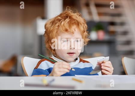 Cute diligent gingerhaired learner of nursery school with crayon of dark green color drawing by desk at lesson during individual work Stock Photo