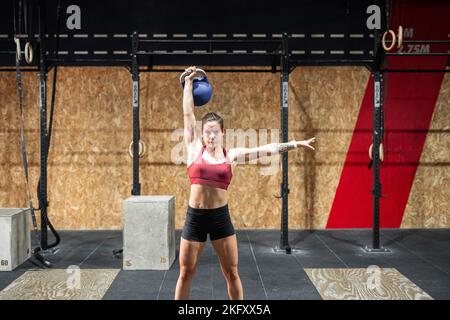 Caucasian girl standing with tattooed arms with her left arm outstretched and her right hand above her head holding a large heavy ball Stock Photo