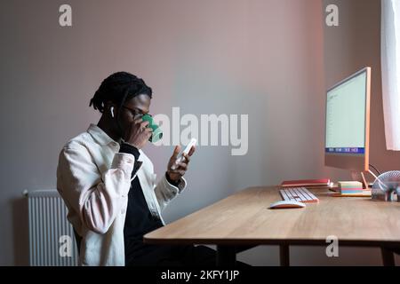 Relaxed carefree man drinking tea sits at desk and using mobile phone sits at table with computer Stock Photo