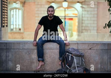 bearded caucasian young man sitting on a wall next to a large backpack smiling at camera barefoot Stock Photo