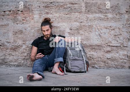 barefoot tired traveler caucasian guy looking and touching his smartphone sitting leaning on his big backpack on the floor by the wall with dirty feet Stock Photo