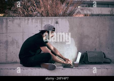 caucasian guy in gray cap black t-shirt sitting on the concrete floor with his legs crossed manipulating his laptop at sunset in the city Stock Photo