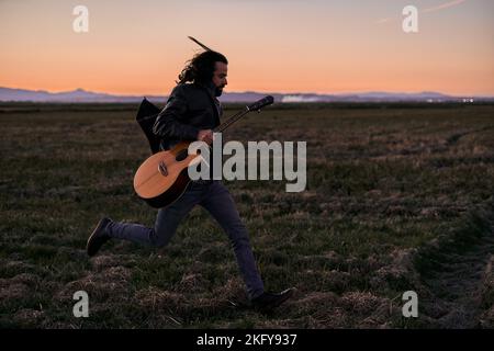 caucasian young man with long hair in fur jacket running through the grass of the field holding a guitar at sunset Stock Photo