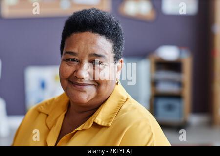 Mature smiling black woman in yellow blouse sitting in front of camera and looking at you against school supplies in classroom Stock Photo