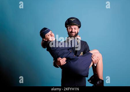 Smiling pilot holding flight attendant in arms portrait, aircrew in professional uniform looking at camera. Happy aviator and stewardess romantic couple studio medium shot Stock Photo