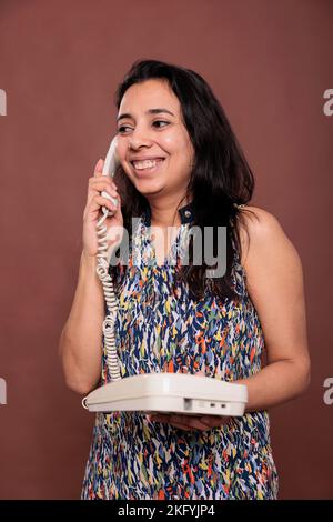 Smiling indian woman answering landline phone call, customer support service, cheerful consultant. Lady chatting, person with positive facial expression holding retro telephone Stock Photo