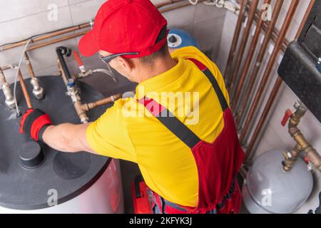 Professional Caucasian Plumber Repairing Tank Water Heater in Boiler Room of Commercial Building. Heating Equipment Maintenance Theme. Stock Photo