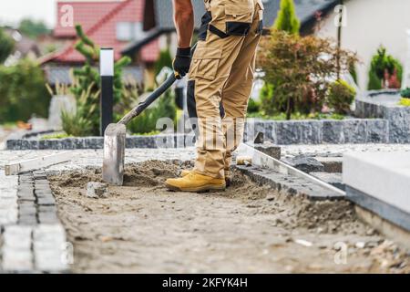 Professional Construction Worker Evenly Spreading Sand with a Shovel Before Laying Paving Bricks. Garden Walkway Building Process. Stock Photo