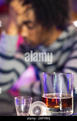 Vertical of drunk african american man holding head at bar, glasses in foreground, selective focus Stock Photo
