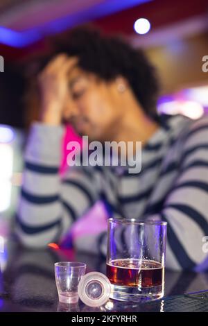 Vertical of drunk african american man holding head at bar, glasses in foreground, selective focus Stock Photo