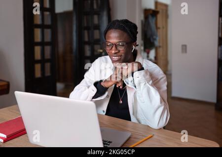 Cheerful inspired African American man smiling looking at laptop screen sits at table in apartment Stock Photo
