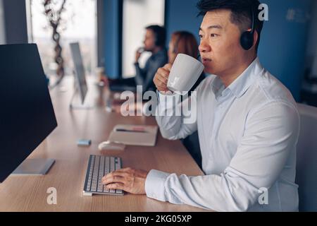 Focused asian businessman drinking coffee while working on computer in modern office Stock Photo