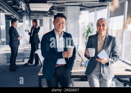 Two happy multiracial colleagues talking and drinking coffee during break time in office Stock Photo
