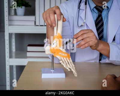 Close-up pen holding by orthopedic doctor man's hand in white coat pointing to foot skeleton, ankle joint anatomy model on desk to explain for patient Stock Photo