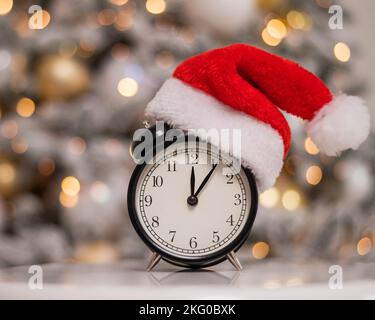 Alarm clock in a santa hat against the background of a christmas tree. Stock Photo