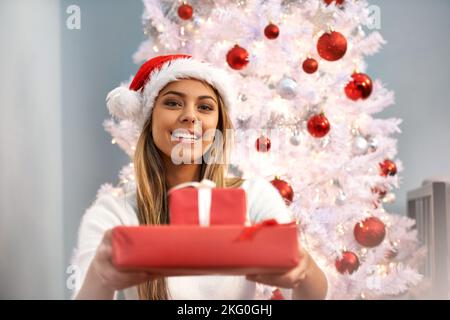 And here is your present. Portrait of a beautiful young woman holding presents while sitting by a Christmas tree. Stock Photo