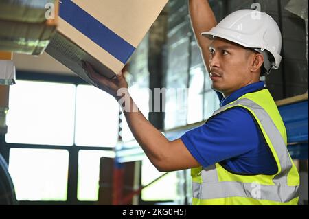 Concentrated millennial Asian male warehouse worker in uniform and hardhat lifting an inventory box, working in warehouse. Stock Photo