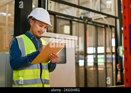 Professional and smart millennial Asian male industrial engineer or warehouse engineer in uniform and white hardhat checking list on his clipboard, wo Stock Photo