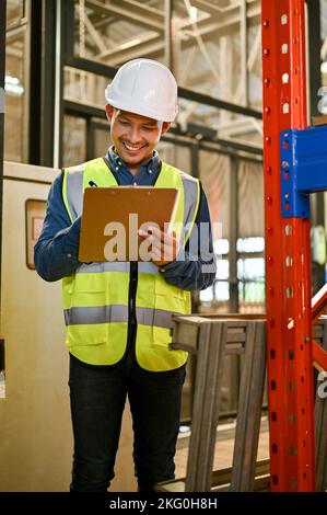 Professional and smart millennial Asian male industrial engineer in uniform and white hardhat checking list on his clipboard, working in factory. Indu Stock Photo
