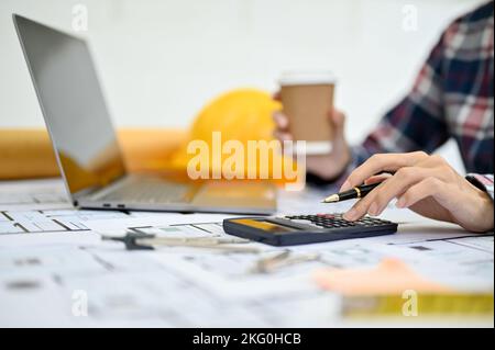 Professional Asian female engineer or architect working on blueprint, using calculator and sipping coffee while working in office. cropped image Stock Photo