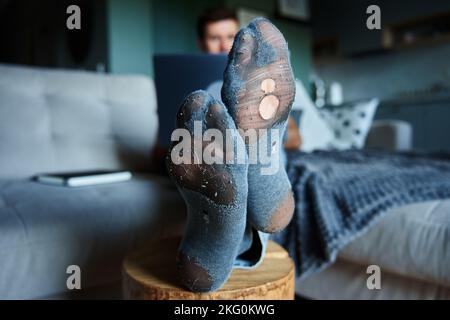 Freelancer work from home. Man in leaky socks using laptop and resting on sofa Stock Photo