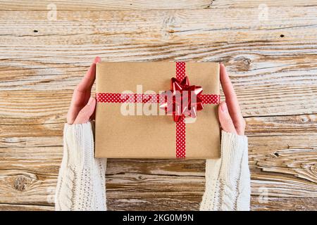 Woman holds gift box wrapped in craft paper with festive ribbon agains wooden background. Female hands holding present for christmas holidays, above view Stock Photo