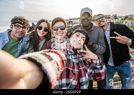 Multiethnic group of friends taking a selfie by the sea - multiracial young people posing together for a group picture standing together seaside - peo Stock Photo