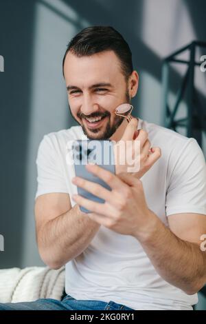 Smiling bearded man using jade face roller for beauty facial massage therapy while looking at the screen of the phone in his hands. Beauty treatment Stock Photo