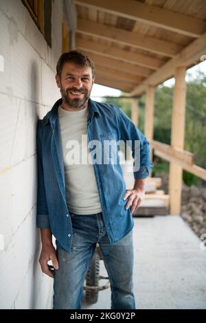 Happy mature man standing in front of his new house under construction, looking at camera. Stock Photo
