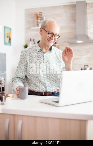 Old man saying hello during video call with family using notebook in kitchen while holding a cup of coffee. Stock Photo