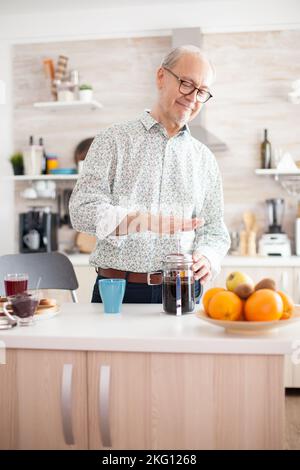 Senior man making coffee using french press during breakfast in kitchen. Elderly person in the morning enjoying fresh brown cafe espresso cup caffeine from vintage mug, filter relax refreshment Stock Photo