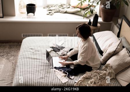 A pregnant woman in a warm sweater is sitting on the bed and buying things for the baby through an online website.The concept of e-commerce, holiday shopping, gift shopping for the birth of a baby. Stock Photo