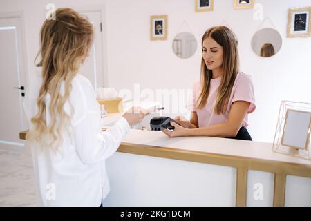 Female customer pay for procedures in beauty studio. Cashier hand holding credit card reader machine while client holding phone for NFC payment Stock Photo