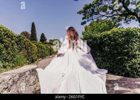 a beautiful woman with long brown hair and long white dress runs along a path along beautiful bushes in the park rear view Stock Photo